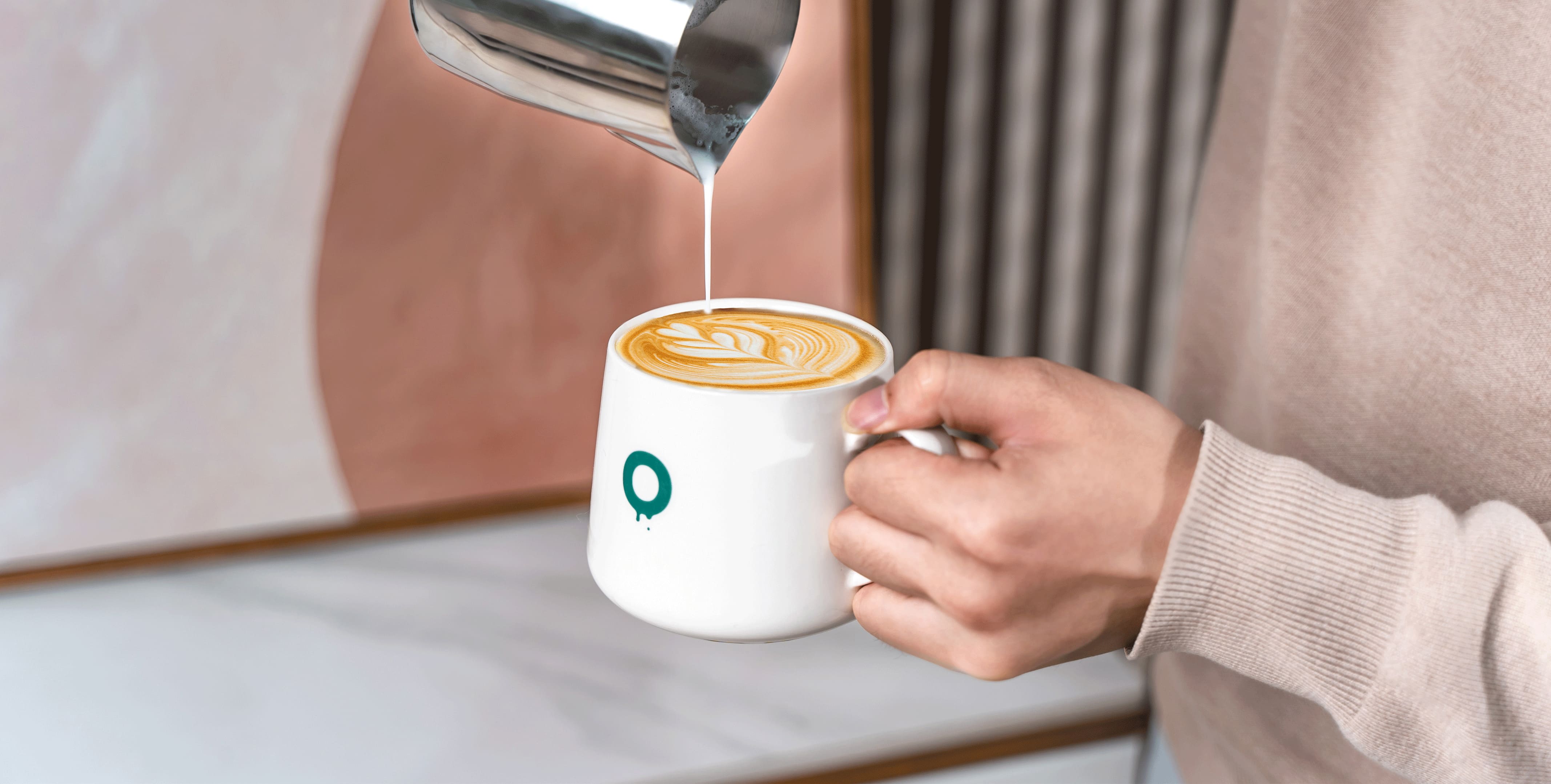 A barista pouring steamed milk into a latte in a white mug with the O logo on it