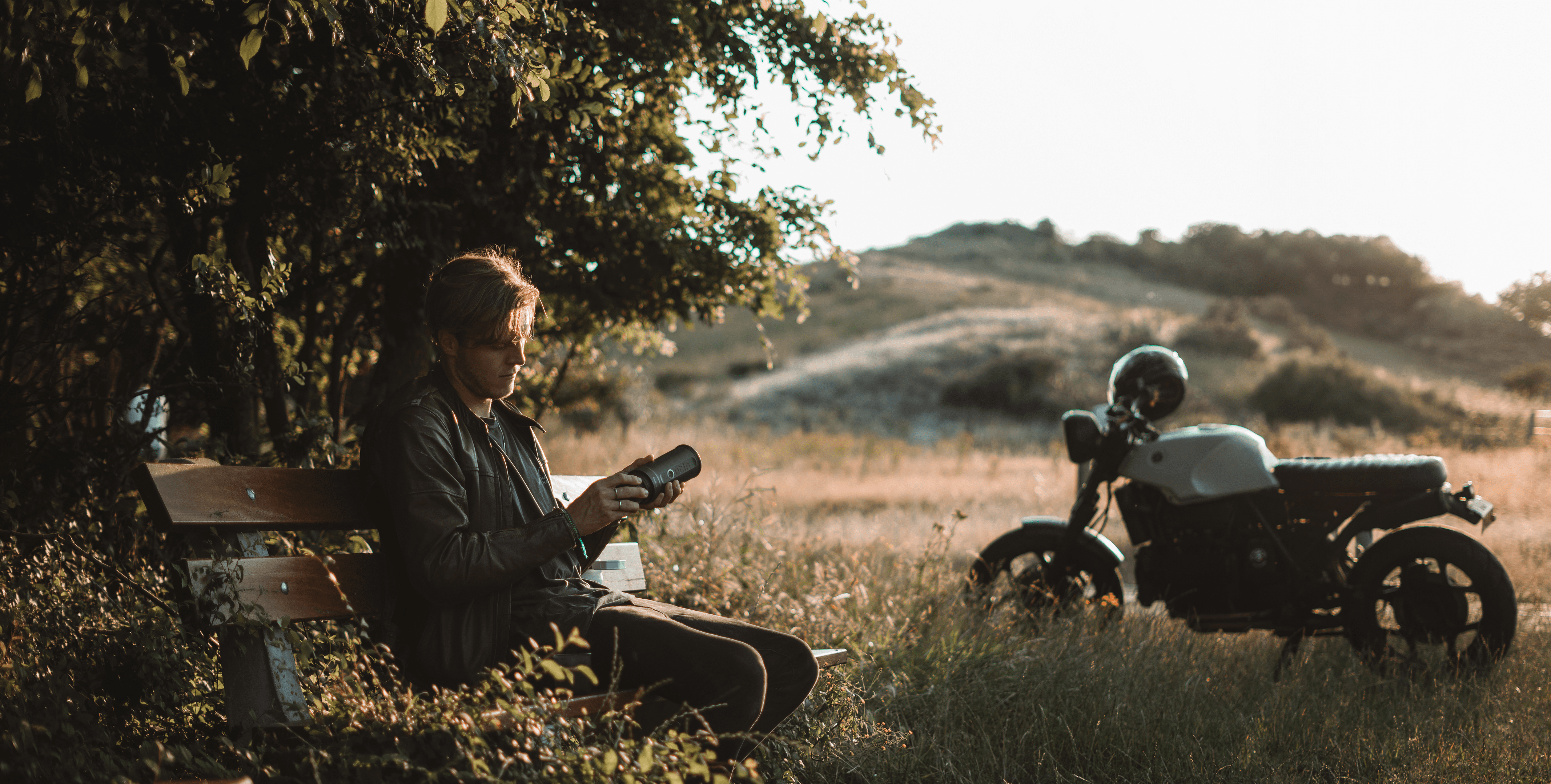 Adventurous motorcyclist enjoys Outin Nano Portable Espresso Machine on countryside bench, vintage motorcycle in background at golden hour